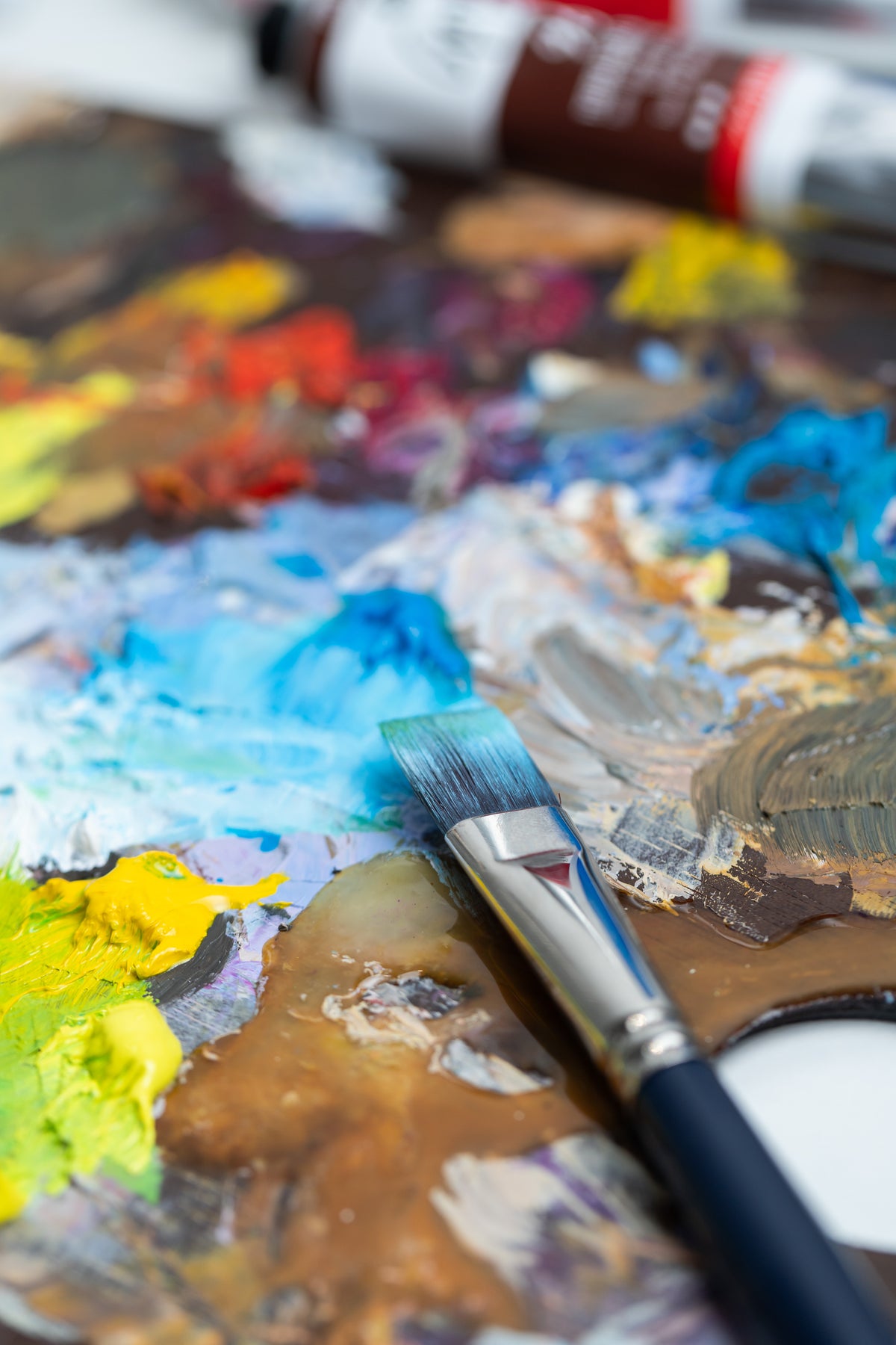 A close-up of a paintbrush resting on a heavily mixed artist palette filled with blue, yellow, brown, white, and red paint, with tubes of paint blurred in the background.