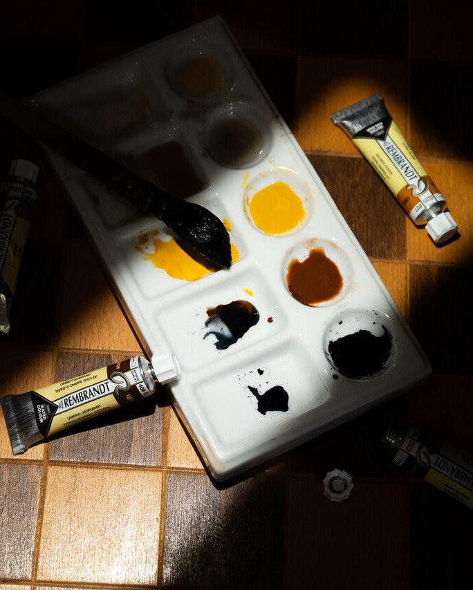 A white paint palette with wells containing yellow, brown, black, and white paint, surrounded by tubes of Winsor & Newton artist paint on a dark wooden checkerboard surface under dramatic spotlighting.