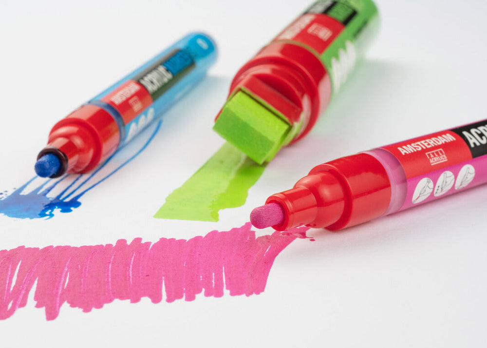 Three acrylic paint markers with colored caps on a white background, each drawing a line in blue, green, and pink.